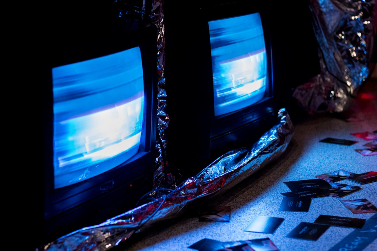 Old CRT televisions displaying blue glitch art at an exhibition with promotional cards scattered on the floor in the foreground.