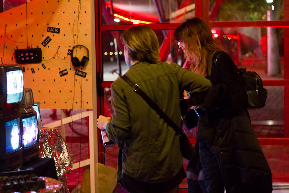 Visitors interacting with a multimedia art installation, which includes CRT TVs displaying visuals, with headphones and other electronic devices mounted on a pegboard, at a glitch art exhibition.