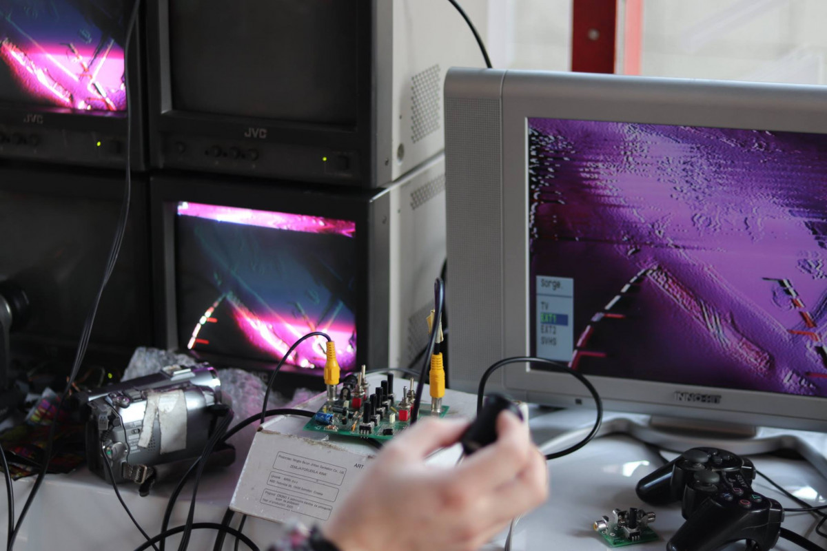 An interactive installation at an exhibition featuring a circuit-bent video glitching setup with multiple CRT monitors displaying distorted, colorful imagery, electronic components, and various cables connected to a central processing unit.