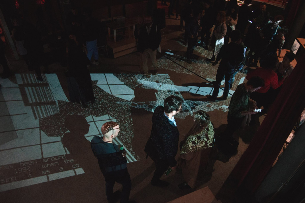 Overhead view of an exhibition space where visitors are cast in silhouette by floor projections of digital glitch art, creating a dynamic and interactive environment.