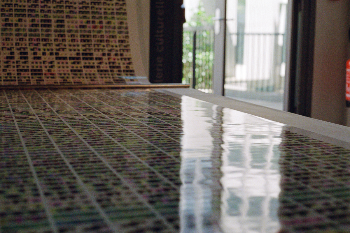 A perspective view of an art exhibition space where a lengthy print is displayed. The print starts from the ceiling and cascades down to eye level, where it extends horizontally across a table, inviting viewers to examine its intricate patterns of densely packed images.