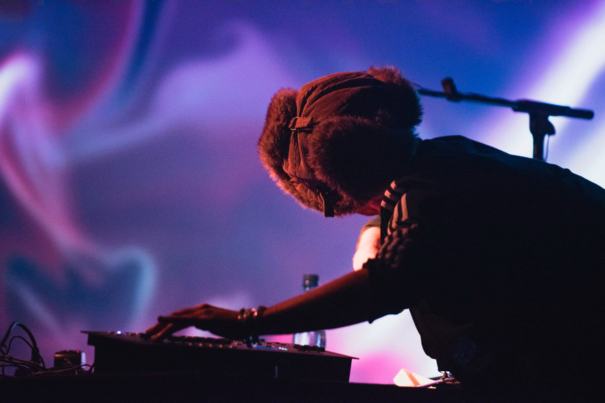 VJ wearing a winter hat adjusting equipment with abstract purple and blue visuals projected in the background, highlighting a creative performance atmosphere